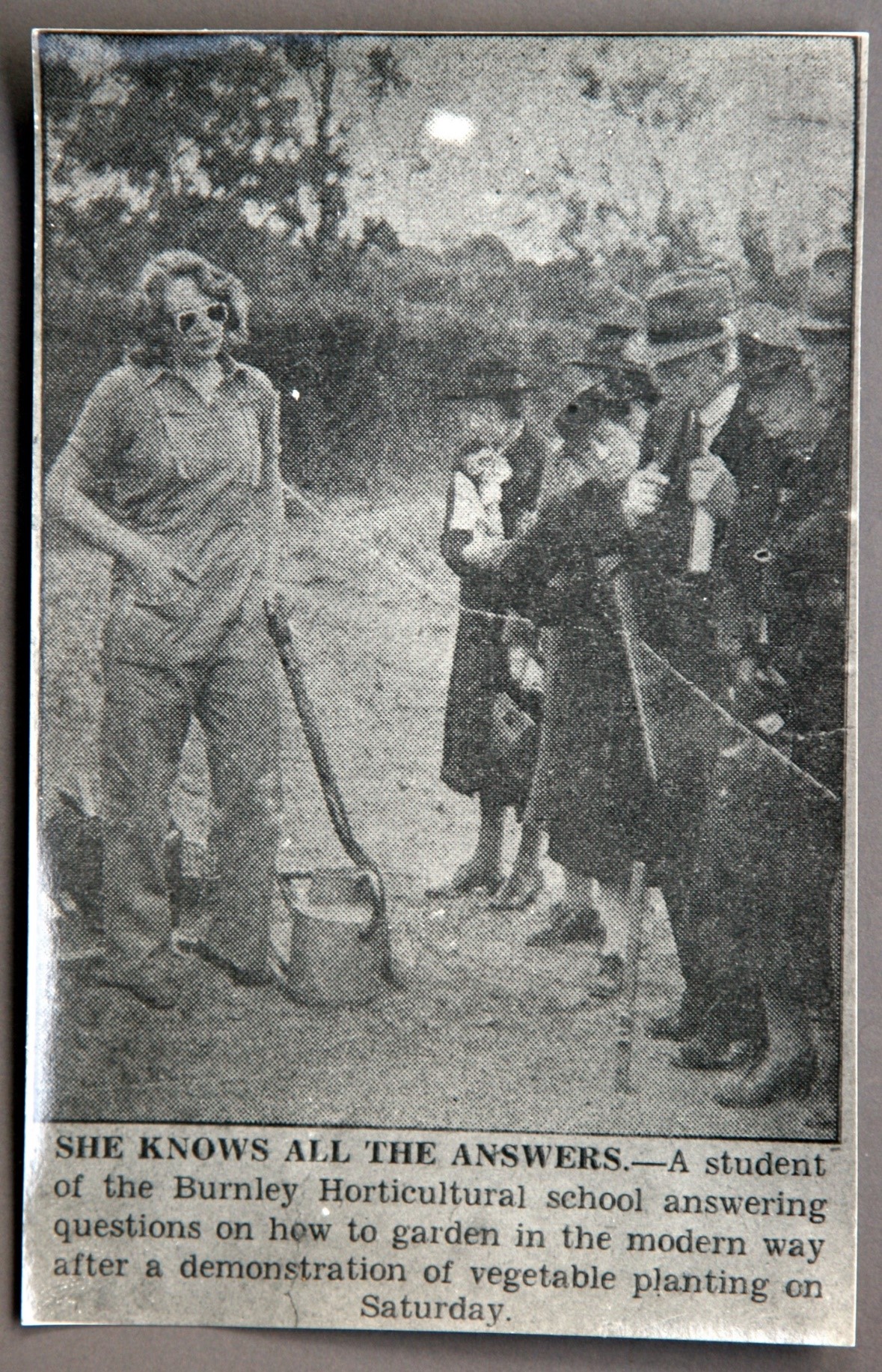 A newspaper clipping of a trendy looking woman in sunglasses and overalls demonstrating to a group of people dressed in long formal coats and hats. Caption reads: "She knows all the answers - A student of the Burnley Horticultural school answering questions on how to garden in the modern way after a demonstration of vegetable planting on Saturday.